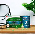 Une boîte de Forté Pharma Expert Cheveux Triple Action, 3x28 comprimés, format 3 mois, dont 50 % propose des compléments de beauté sur un plateau en bois à côté d'une plante, avec des comprimés verts éparpillés devant.