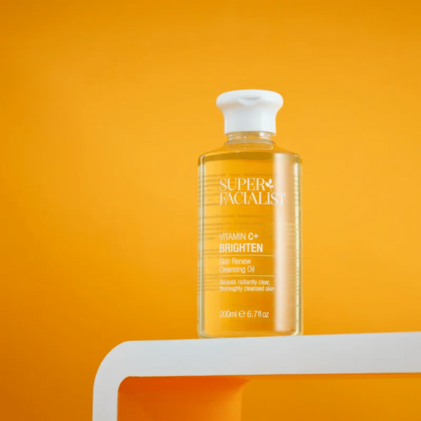 Un flacon de Super Facialist Vitamin C+ Brighten Huile Démaquillante Purifiante Peau Lumineuse 200 ml sur étagère blanche, sur fond orange, disponible chez Univers Cosmetix.