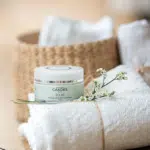 Un pot de CANDES Masque Exfoliant Clarifiant 50 ml posé sur des serviettes blanches roulées, décorées de petites fleurs blanches, un panier en osier en arrière-plan.