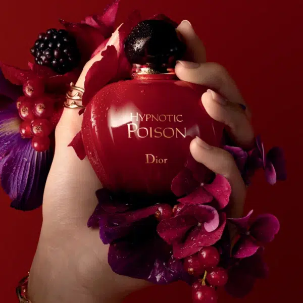 Main tenant le flacon DIOR Hypnotic Poison Eau de Toilette pour femme 30 ml entouré de fleurs violettes et rouges sur fond rouge, dégageant un charme rappelant Miss DiorParfum.