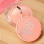 Un contenant rose de Coffret Gel à Sourcils Liftant Essence Cosmetics 12 g, accompagné d'un pinceau inclus, posé sur une surface beige texturée.