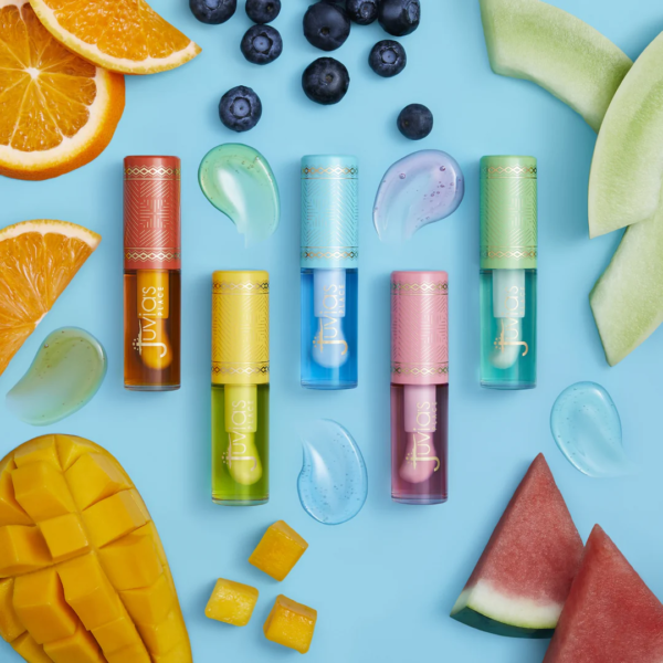 Six tubes vibrants d'huiles à lèvres Juvias Magic Lip Oils 1 g disposés sur un fond bleu, entourés de fruits frais et d'échantillons de gloss.