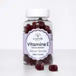 Une bouteille de gummies LASHILE BEAUTY Vitamine E Agit contre le stress oxydatif 60 pièces avec quelques-uns éparpillés devant. L'étiquette indique « 60 pièces, vegan » et met en évidence son avantage : « Agit contre le stress oxydatif.