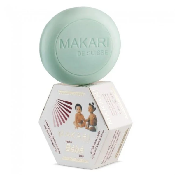 Un savon rond vert étiqueté « Makari de Suisse » repose sur une boîte hexagonale indiquant « Makari Bébé Savon au Calendula et Beurre de Karité 155 g », enrichi au beurre de karité et au calendula et orné d'images de bébé.