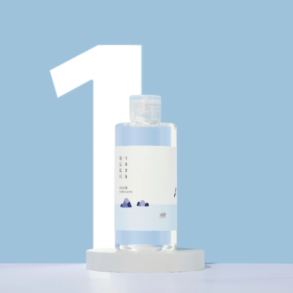 Un flacon transparent Round Lab1025 Dokdo Tonique Exfoliant 100 ml sur un socle blanc, avec un grand chiffre "1" en arrière-plan sur fond bleu.