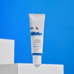 Un tube de Round Lab Birch Moisturizing Crème Spf50 aux Bouleaux Rafraîchissante 50 ml avec un emballage blanc et bleu, posé sur des blocs blancs sur fond bleu vif.