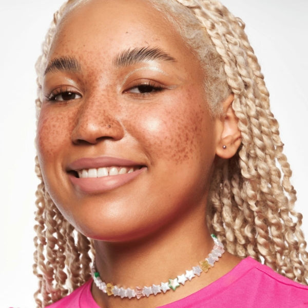 Une personne souriante avec des tresses blondes claires, des taches de rousseur, portant un collier étoile et une chemise rose vif, mettant en valeur ses joues roses avec Essence Cosmetics Blush Liquide Baby Got Blush. dakar, Sénégal