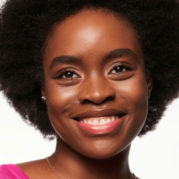 Une personne souriante avec une coiffure afro et un haut rose vif, mettant en valeur sans effort sa beauté naturelle sur un fond blanc uni, une combinaison parfaite pour l'éclat lumineux du Blush Liquide Baby Got Blush d'Essence Cosmetics. dakar, Sénégal