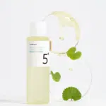 Un flacon de Numbuzin Tonique Clarifiant Anti Taches No.5+ Vitamin C Boosting 200 ml, réputé pour son mélange similaire à Some By Mi Yuja Niacin, se trouve à côté d'une assiette circulaire contenant du liquide et des feuilles vertes.