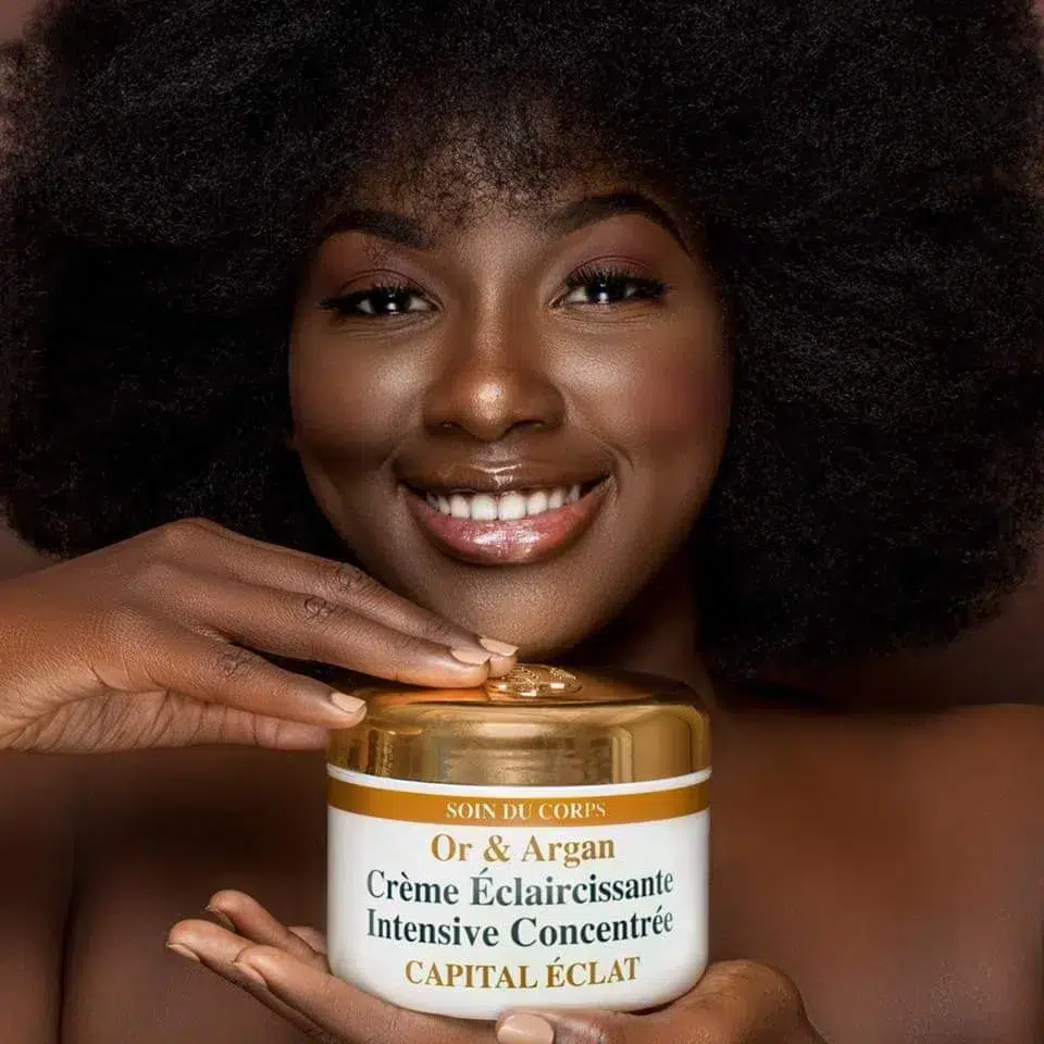 Femme souriante avec une coupe afro tenant un pot de HT26 Or et Argan Crème Corps Éclaircissante 500 ml. dakar