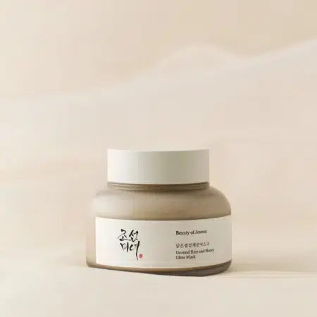 Un pot de Masque Éclat au Riz Moulu et au Miel 150 ml de Beauty of Joseon repose sur un fond doux et neutre. Le pot beige, arborant une étiquette coréenne, respire l'élégance minimaliste et la beauté intemporelle. Dakar