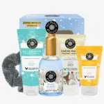 Le Coffret Nuit Étoilée Musc Blanc de LES PETITS PLAISIRS comprenant un chouchou gris, un gel douche, une crème pour les mains, une lotion pour le corps et un flacon de parfum est présenté devant une boîte décorative. Dakar