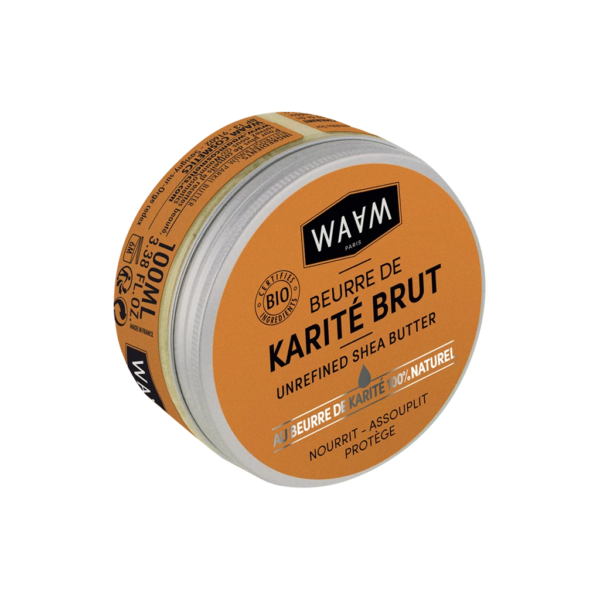 Une boîte ronde de beurre de karité non raffiné de marque WAAM avec une étiquette orange et un texte noir et blanc. L'étiquette met en avant la certification biologique et les propriétés nourrissantes, adoucissantes et protectrices. Le couvercle est légèrement ouvert, laissant apparaître une substance blanche. Dakar