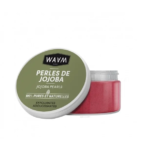 Un pot de Perles de Jojoba WAAM avec le couvercle partiellement ouvert. Le couvercle est vert avec du texte et le récipient contient des perles exfoliantes roses étiquetées comme 100% pures et naturelles. Dakar