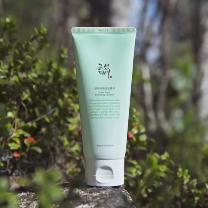 Un tube de nettoyant rafraîchissant à la prune verte Beauty of Joseon est posé sur un rocher en plein air, entouré d'une végétation floue. L'emballage vert présente des textes en anglais et en coréen. La lumière du soleil met le produit en valeur, soulignant le cadre naturel. Dakar
