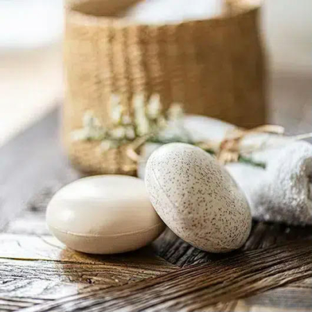 Deux savons ovales, l'un lisse, l'autre moucheté, reposent sur une surface tissée. Une serviette blanche roulée et un panier en osier beige orné de fleurs blanches se détachent en arrière-plan flou, créant une ambiance de spa. Dakar