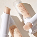 Trois tubes de baume à lèvres vegan au thé kombucha sont exposés, l'un d'eux ouvert laissant apparaître le baume. L'emballage blanc présente un étiquetage minimaliste, projetant des ombres douces sur le fond clair. Dakar