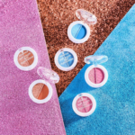 Six boîtiers de fards à paupières Essence Soft Touch sont disposés sur une surface texturée pailletée rose, bleue et cuivrée. Leurs teintes varient, notamment le bleu, le marron et le rose. Chaque boîtier est ouvert, laissant apparaître les couleurs des fards à paupières. Dakar