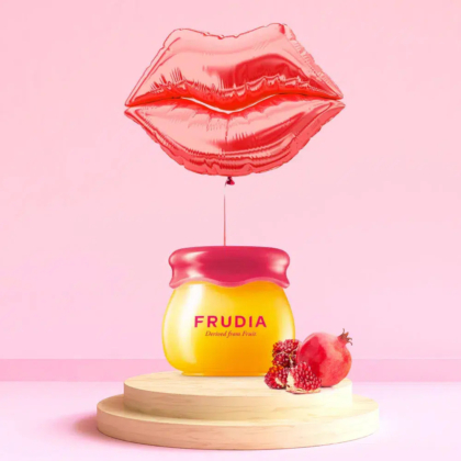 Un petit pot de crème de soin Frudia posé sur une plateforme en bois, accompagné d'une grenade coupée en deux et de ses graines. Au-dessus, un ballon rouge en forme de lèvres flotte sur un fond rose. Dakar