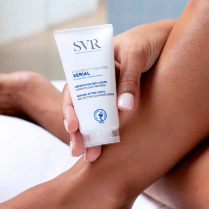 Une personne applique la crème SVR Xerial sur son pied. La crème est présentée dans un tube blanc avec un texte bleu et noir, favorisant l'hydratation et la protection du pied. La main de la personne tient le tube, et l'attention est portée sur le pied et la crème. Dakar